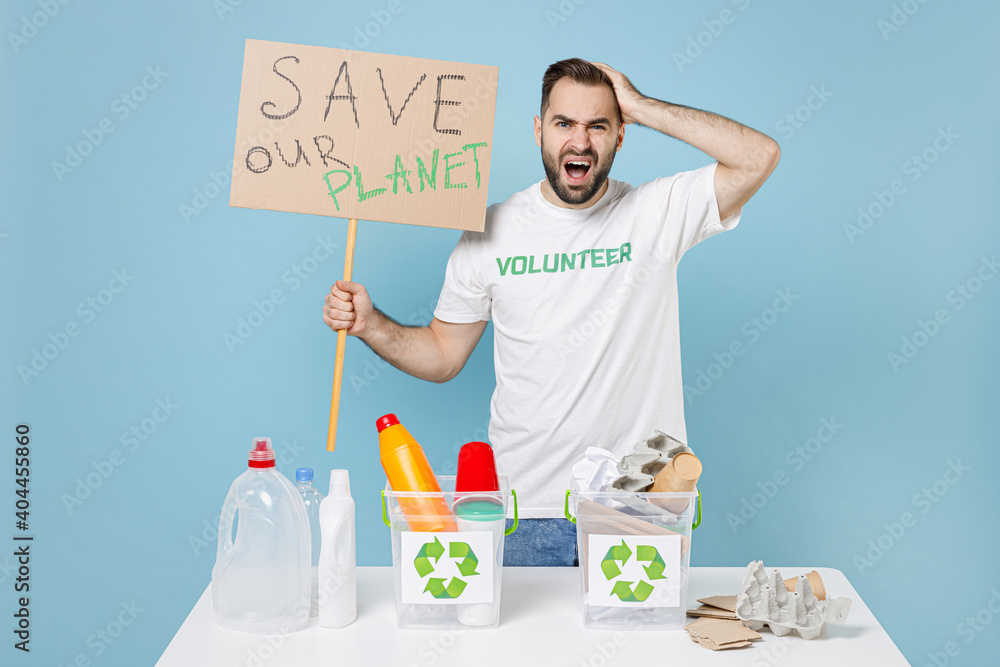 Worried man in volunteer tshirt stand near recycling stations sorting plastic paper trash hold nameplate save planet hand on head isolated on blue background. Voluntary free work assistance concept. Worried man in volunteer tshirt stand near recycling stations sorting plastic paper trash hold nameplate save planet hand on head isolated on blue background. Voluntary free work assistance concept.