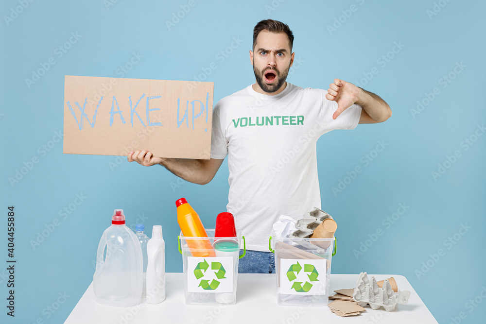 Shocked man in volunteer tshirt stand near recycling stations sorting plastic paper trash hold nameplate wake up showing thumb down isolated on blue background. Voluntary free work assistance concept. Shocked man in volunteer tshirt stand near recycling stations sorting plastic paper trash hold nameplate wake up showing thumb down isolated on blue background. Voluntary free work assistance concept.