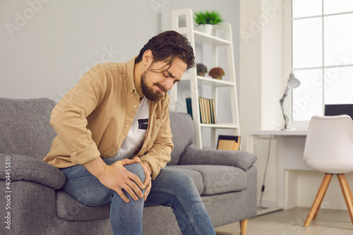 Unhappy man sitting on sofa at home and touching hurt knee with grimace of pain and suffering. Young guy with rheumatism, arthritis or osteoarthritis feeling intense severe pain in knee joint © Studio Romantic