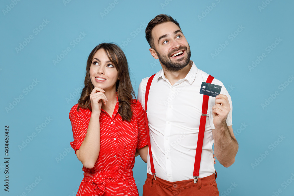 Pretty pensive young couple two friends man woman wearing white red clothes hold credit bank card looking aside up isolated on pastel blue color background studio. St. Valentine's Day holiday concept. Pretty pensive young couple two friends man woman wearing white red clothes hold credit bank card looking aside up isolated on pastel blue color background studio. St. Valentine's Day holiday concept.