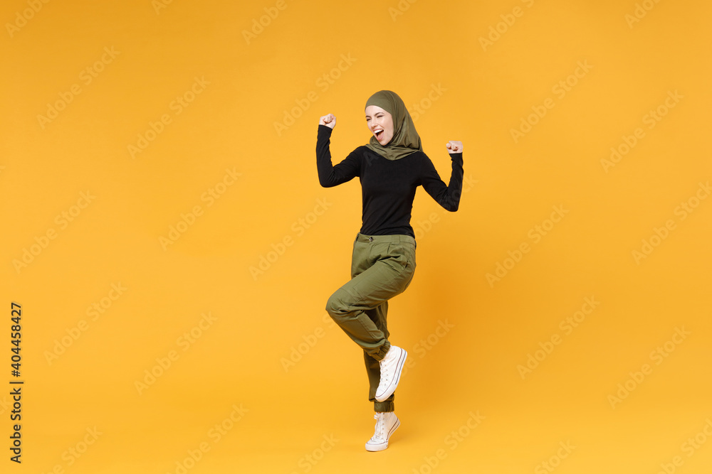Full length joyful arabian muslim woman in hijab black green clothes clenching fists like winner celebrating say yes isolated on yellow background studio portrait. People religious lifestyle concept.
