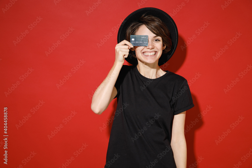 Smiling funny young brunette woman 20s years old wearing casual basic black t-shirt hat stand covering eye with credit bank card looking camera isolated on bright red color background studio portrait. Smiling funny young brunette woman 20s years old wearing casual basic black t-shirt hat stand covering eye with credit bank card looking camera isolated on bright red color background studio portrait.