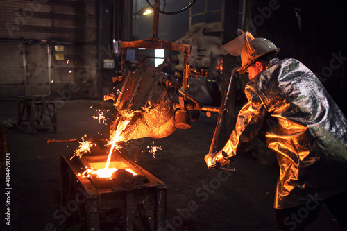 Fényképezés Foundry worker in protective suit and hardhat filling mold with hot molten iron to make parts for industry