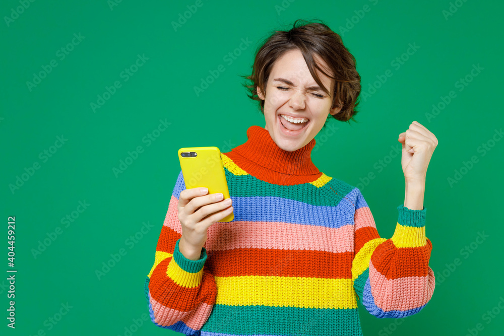Happy young brunette woman 20s wearing basic casual colorful sweater standing using mobile cell phone typing sms message doing winner gesture isolated on bright green color background studio portrait. Happy young brunette woman 20s wearing basic casual colorful sweater standing using mobile cell phone typing sms message doing winner gesture isolated on bright green color background studio portrait.