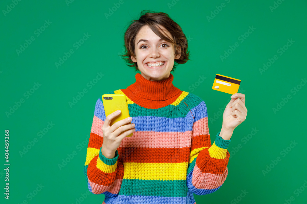 Cheerful young brunette woman 20s years old wearing basic casual colorful sweater standing using mobile cell phone hold credit bank card isolated on bright green color background studio portrait. Cheerful young brunette woman 20s years old wearing basic casual colorful sweater standing using mobile cell phone hold credit bank card isolated on bright green color background studio portrait.