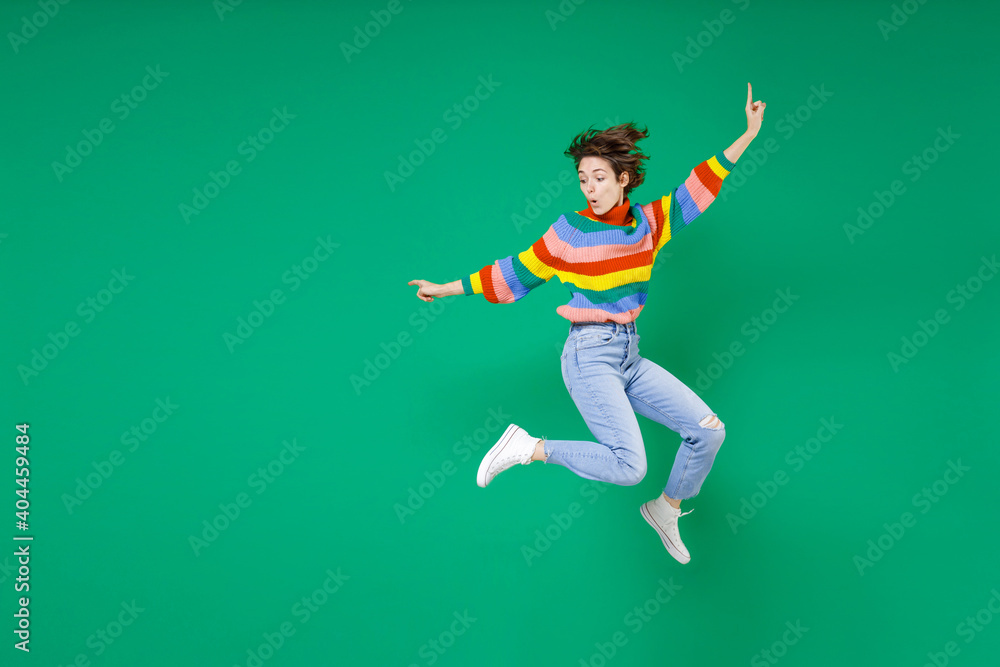 Full length side view of amazed young brunette woman 20s years old in casual colorful sweater jumping spreading hands pointing fingers aside isolated on bright green color background studio portrait.