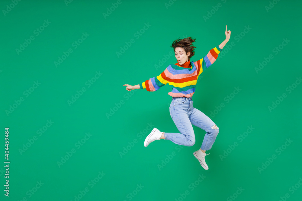 Full length side view of amazed young brunette woman 20s years old in casual colorful sweater jumping spreading hands pointing fingers aside isolated on bright green color background studio portrait. Full length side view of amazed young brunette woman 20s years old in casual colorful sweater jumping spreading hands pointing fingers aside isolated on bright green color background studio portrait.