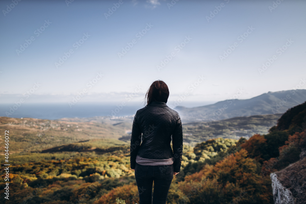 caucasian woman in dark jeans and a dark jacket stands on the edge of a cliff, and looks at the landscape of an autumn valley going to the sea against a blue sky.
