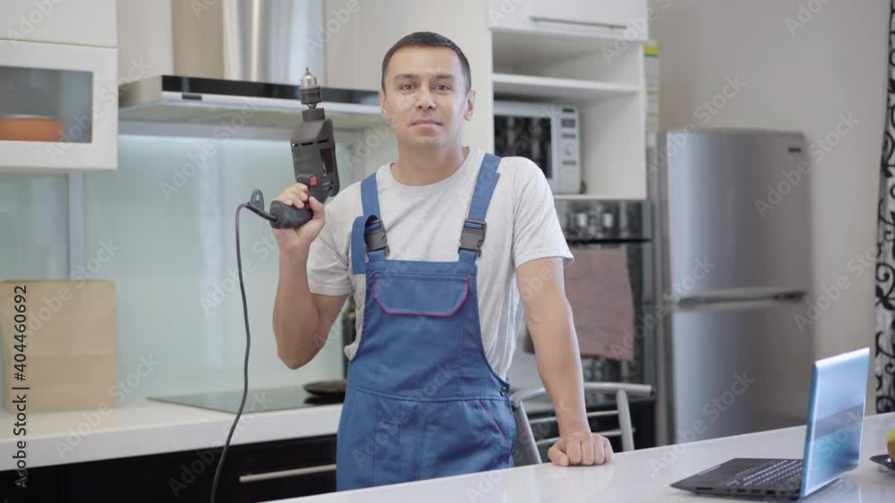 Portrait of confident professional service man posing with electric drill in apartment. Young smiling Caucasian master standing in flat with repair tool and smiling. Fixing and repairing concept. Portrait of confident professional service man posing with electric drill in apartment. Young smiling Caucasian master standing in flat with repair tool and smiling. Fixing and repairing concept.