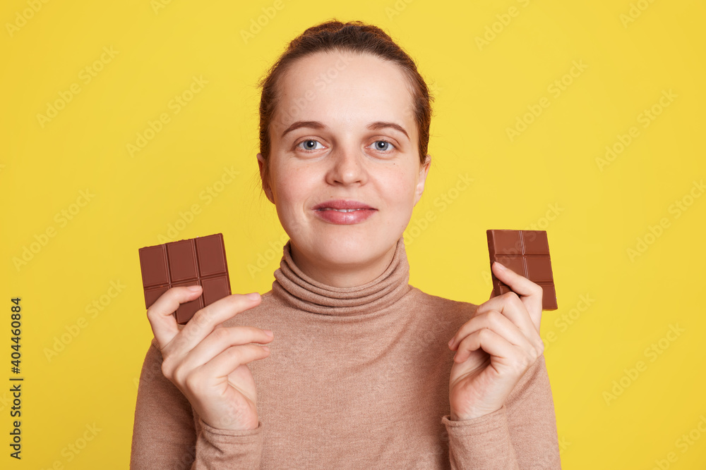 Pregnant woman holding two bars of chocolate in both hands and looking directly at camera with satisfied facial expression, posing isolated over yellow background, wants to eat junk food. Pregnant woman holding two bars of chocolate in both hands and looking directly at camera with satisfied facial expression, posing isolated over yellow background, wants to eat junk food.