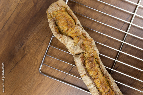 One baguette on a wooden background. A crispy white baguette lies on a wire rack on a dark textured wooden table. View from above.