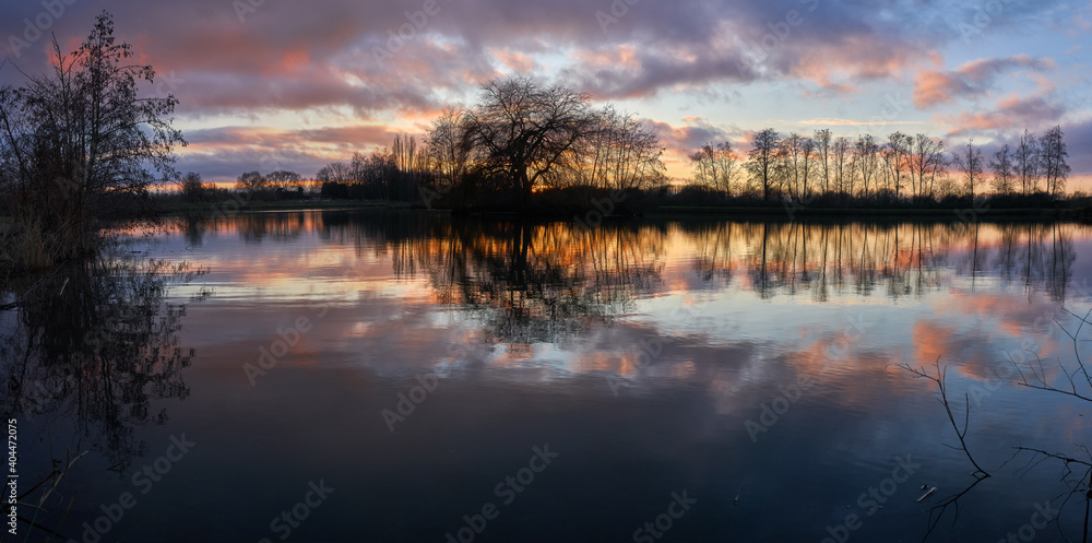 Fototapeta premium scenic panorama of a colorful sunset sky and a row of leafless trees reflecting in a pond in Käseburg (city of Brake Unterweser, Germany)