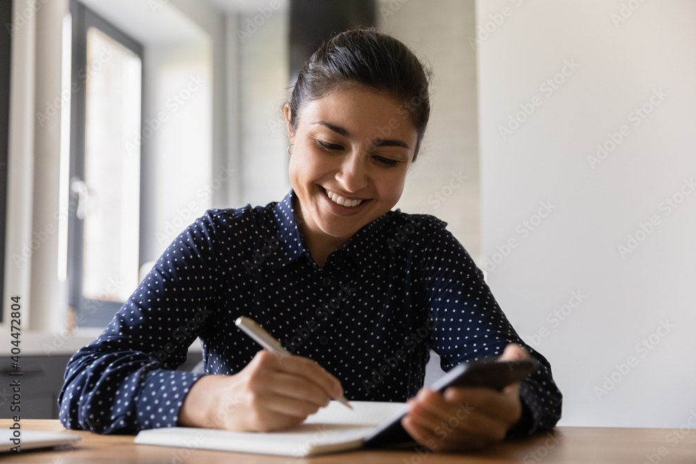 Smiling millennial Indian woman sit at desk make notes studying using modern smartphone. Happy young female write in notebook plan or list looking at cellphone screen. Time management concept. Smiling millennial Indian woman sit at desk make notes studying using modern smartphone. Happy young female write in notebook plan or list looking at cellphone screen. Time management concept.