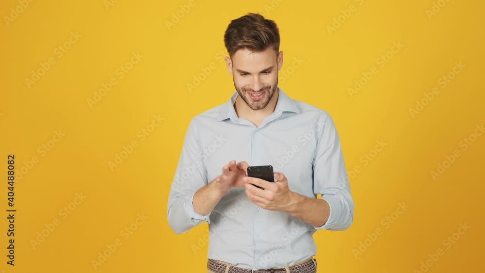 A smiling young man is using his smartphone standing isolated over a yellow background in the studio A smiling young man is using his smartphone standing isolated over a yellow background in the studio