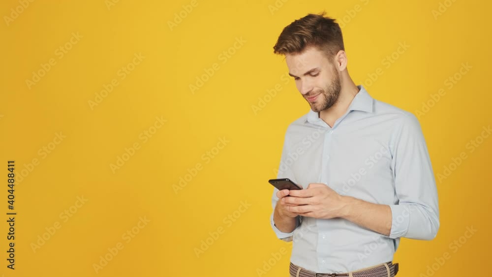 A pleased young man is using his smartphone standing near free space isolated over a yellow background in the studio A pleased young man is using his smartphone standing near free space isolated over a yellow background in the studio