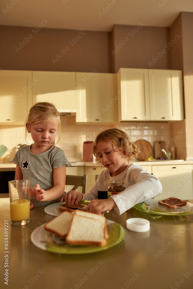 Cute brother spreading chocolate nut butter on toasted bread while preparing breakfast for his little sister and himself in the kitchen