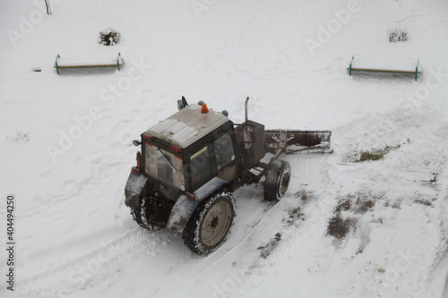 Old wheeled tractor with a shovel removes snow in a pile on the Park after a snowfall on a winter day top back side view on benches background, city utilities