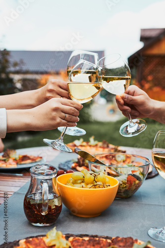 Friends making toast during summer picnic outdoor dinner in a home garden. Close up of people holding wine glasses with white wine over table with pizza, salads and fruits. Dinner in a orchard