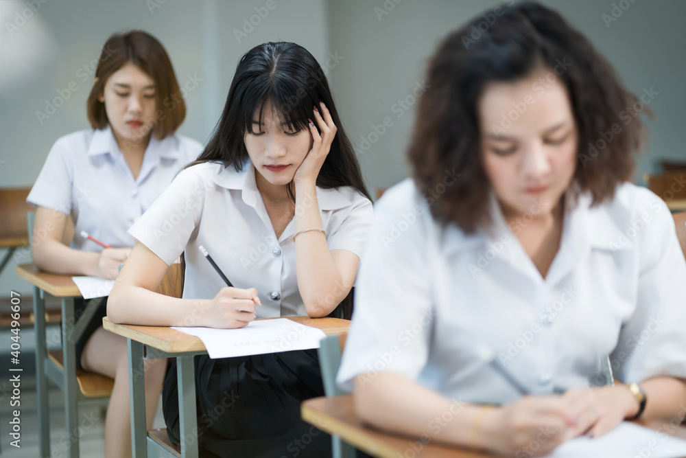 Selective focus of the teen college students sitting on lecture chair in classroom write on examination paper answer sheet in doing the final examination test. Female students in the student uniform. Selective focus of the teen college students sitting on lecture chair in classroom write on examination paper answer sheet in doing the final examination test. Female students in the student uniform.