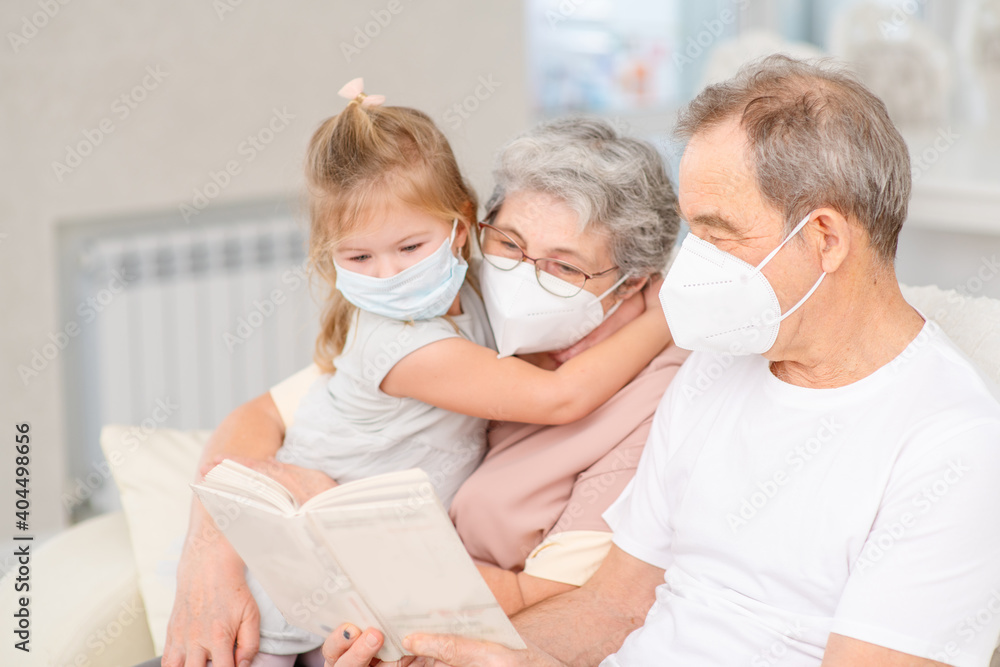 An elderly couple with medical masks are sitting on the couch at home and reading a book to their granddaughter with a medical mask on their face. Ways not to get bored during lockdown and develop whi An elderly couple with medical masks are sitting on the couch at home and reading a book to their granddaughter with a medical mask on their face. Ways not to get bored during lockdown and develop whi