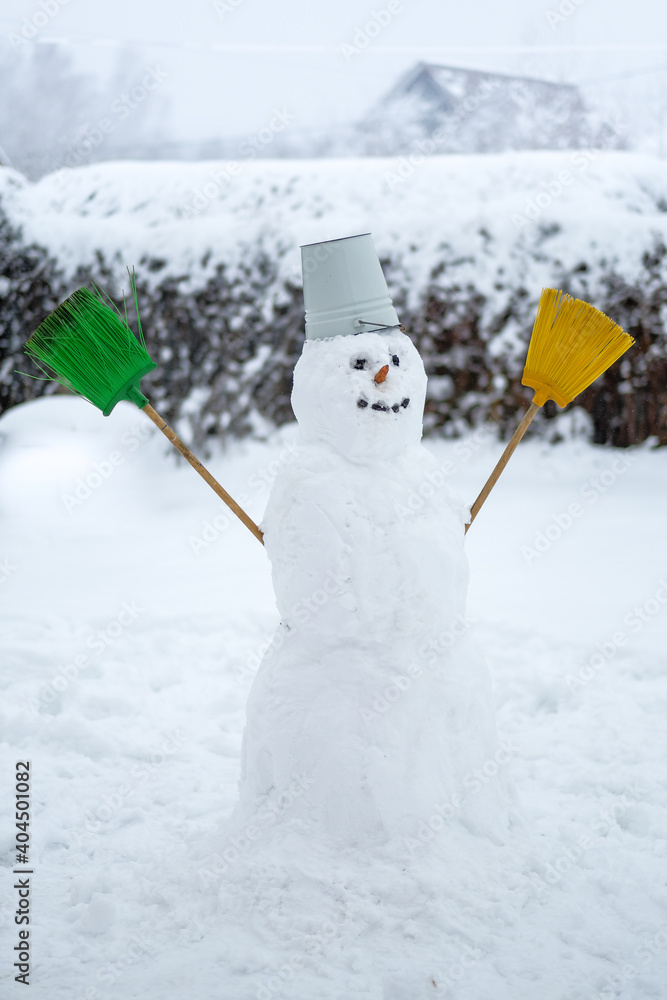 Obraz premium Portrait of a happy white snowman with an orange carrot nose, black embers for eyes, brooms for hands and a bucket on his head. Christmas outdoor fun in winter. Blurred background.