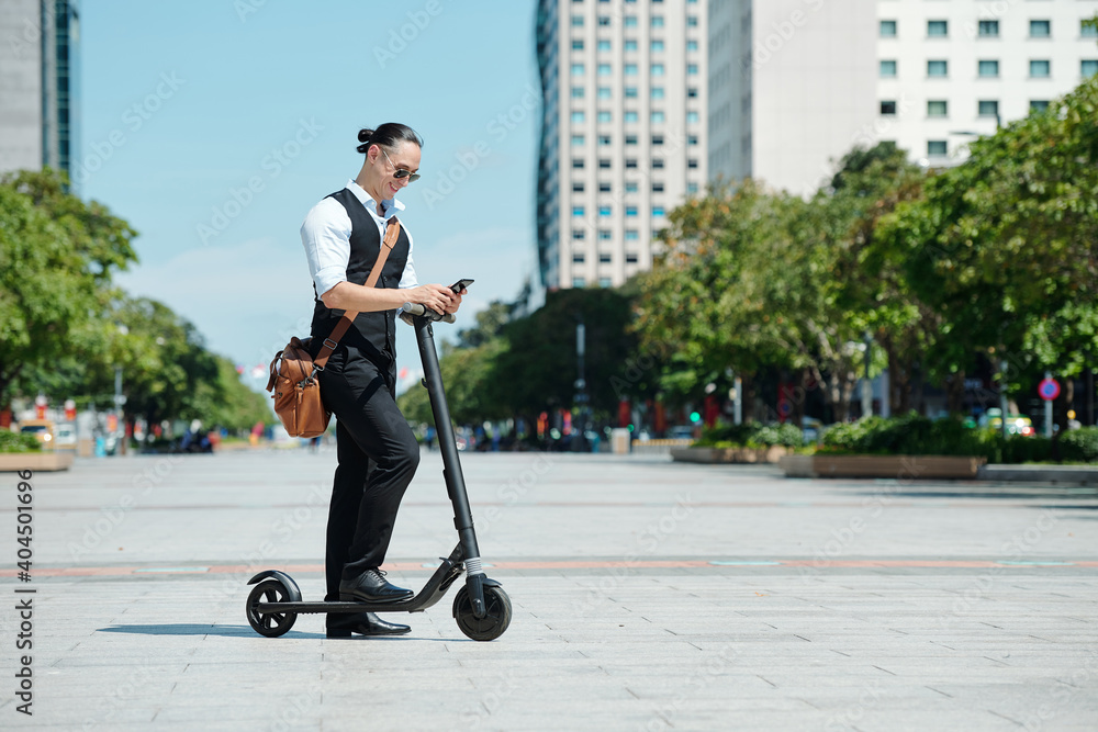 Smiling elegant businessman with electric scooter standing outdoors and reading text messages on smartphone Smiling elegant businessman with electric scooter standing outdoors and reading text messages on smartphone