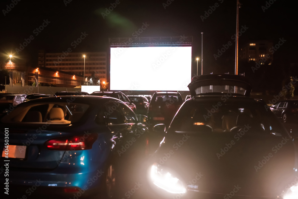 The best place to go. Many cars parked in front of a big white screen to watch movies or films sitting inside the car at drive in cinema in the evening - obrazy, fototapety, plakaty 