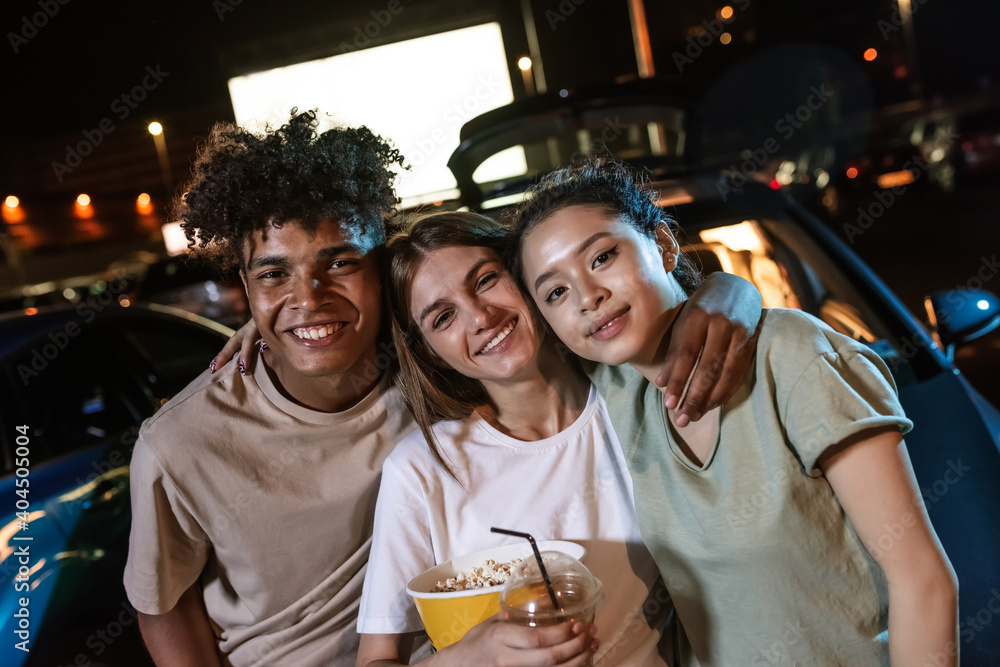 Portrait of three happy diverse young friends smiling at camera while posing together in front of a big screen, ready to watch a movie in an open air cinema