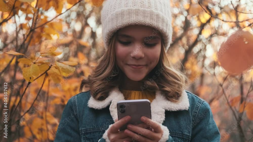 A happy young blonde girl is using her smartphone while standing in the park outside A happy young blonde girl is using her smartphone while standing in the park outside