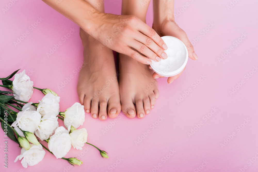 cropped view of barefoot woman holding cosmetic cream near white eustoma flowers on pink background