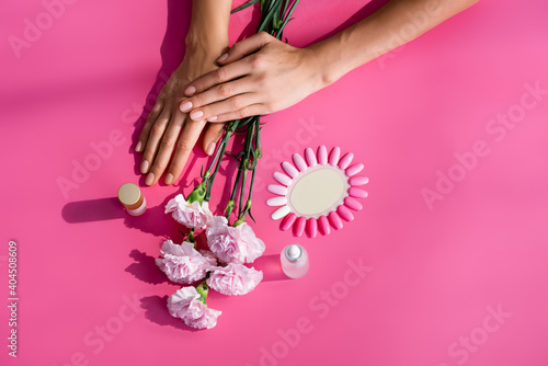 top view of female hands near palette of false nails, carnation flowers, and bottles of cuticle remover and enamel on pink background