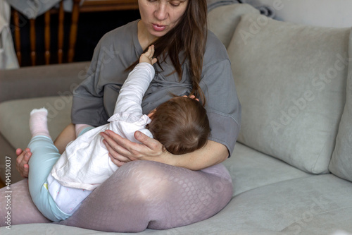 Tired mother breastfeeding her baby daughter while being at home. Beautiful young woman lactating her toddler baby on couch at home, copy space. Family, breastfeeding and motherhood concept. photo