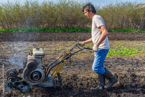 Man in wellingtons with cultivator ploughs ground in sunny day. Land cultivation, soil tillage. Spring work in garden. Gardening concept