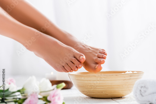 groomed female feet near bowl and flowers on white surface, blurred foreground