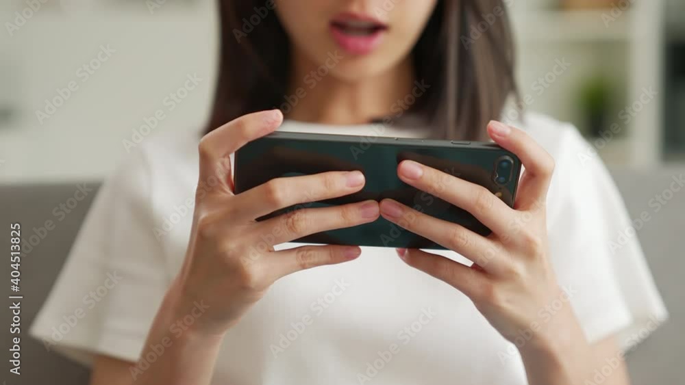 Excited young Asian woman playing games on smartphone in the living room at home. Excited young Asian woman playing games on smartphone in the living room at home.
