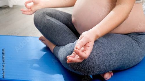 CLoseup of pregnant woman folding her fingers and sitting in lotus yoga pose on fitness mat at big window. Concept of healthy lifestyle, healthcare and sports during pregnancy