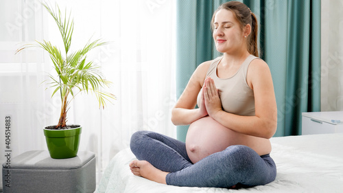 Beautiful smiling pregnant woman breathing deeply and meditating in lotus asana on bed against big window. Concept of healthy lifestyle, healthcare and sports during pregnancy
