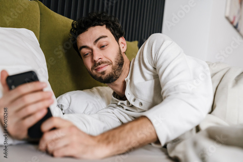 Pleased handsome man using mobile phone while lying in bed after sleep