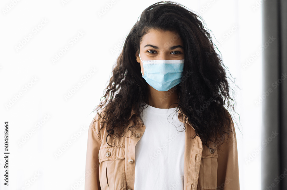 Prevention measures. African american pretty brunette woman in casual wear standing indoors with protective medical mask on her face and looking directly into the camera Prevention measures. African american pretty brunette woman in casual wear standing indoors with protective medical mask on her face and looking directly into the camera
