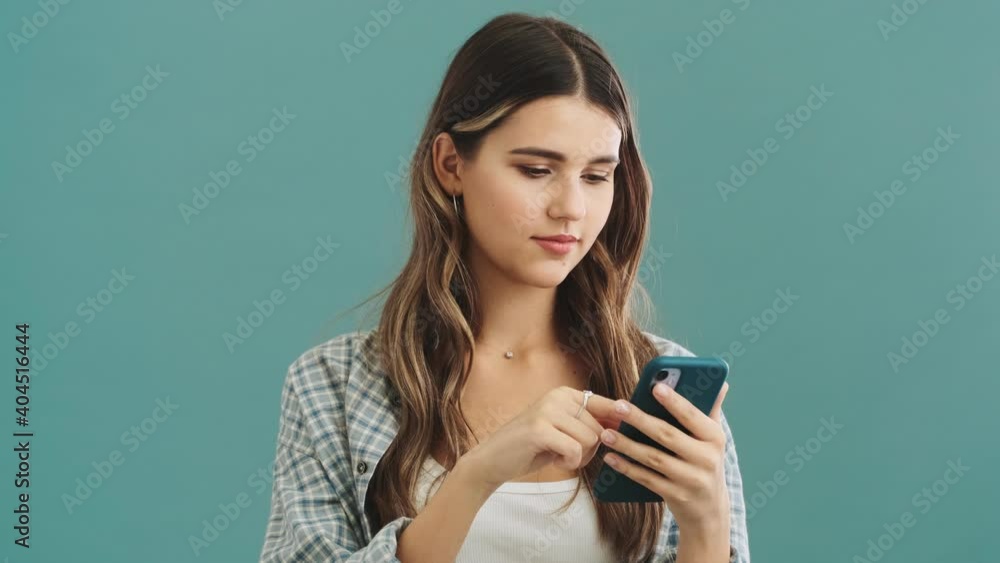 A pleased woman is choosing something while typing on her smartphone isolated over blue background A pleased woman is choosing something while typing on her smartphone isolated over blue background