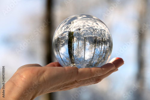 glänzende Glaskugel liegt auf offener Handfläche, verschneiter Wald spiegelt sich darin.