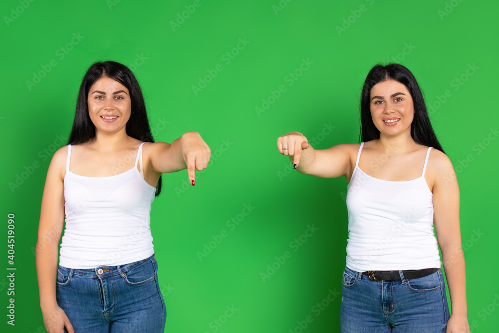 Twins in identical clothes point down with their index finger. Green background and empty space. Twins in identical clothes point down with their index finger. Green background and empty space.