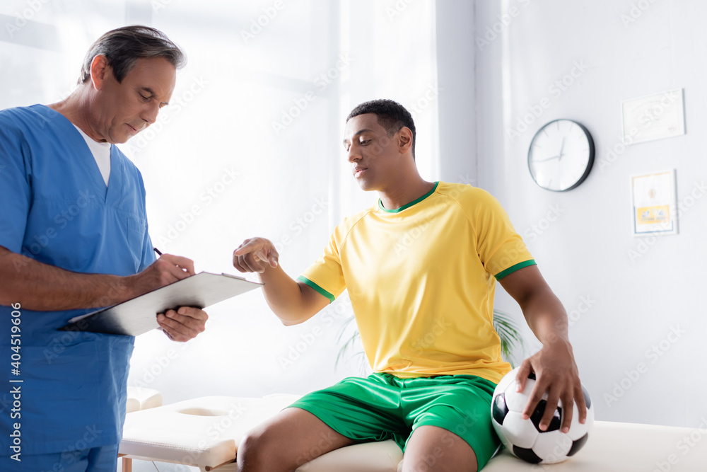 Obraz premium doctor writing prescription near injured african american football player pointing with finger in clinic