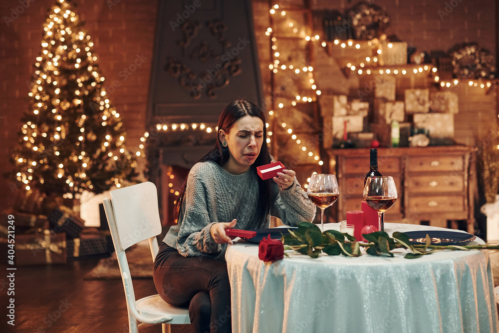 Young disappointed brunette sits by the table in restaurant and holds red box with gift Young disappointed brunette sits by the table in restaurant and holds red box with gift