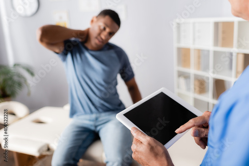 mature doctor using digital tablet near african american patient suffering from neck pain on blurred background