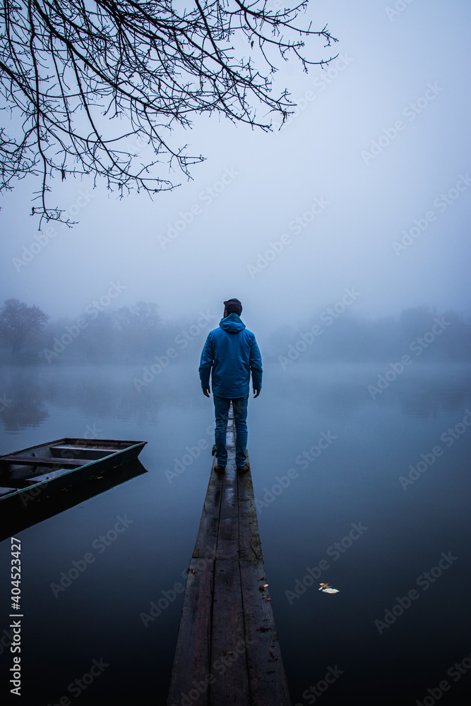 man standing on a pier with a boat at a misty lake early in the morning  
