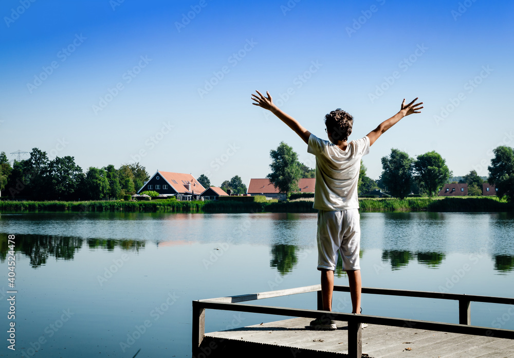 Netherlands, Goes. Dutch countryside lake landscape: reeds alternating with wooden piers. A Caucasian boy is inspired by nature and raises his arms to the sky.
