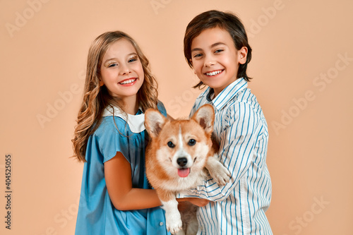 Cute pretty girl with curls in a blue dress and a boy in a shirt hug a funny dog ​​with a bow on his neck isolated on a powdery peach background.Valentine's Day.