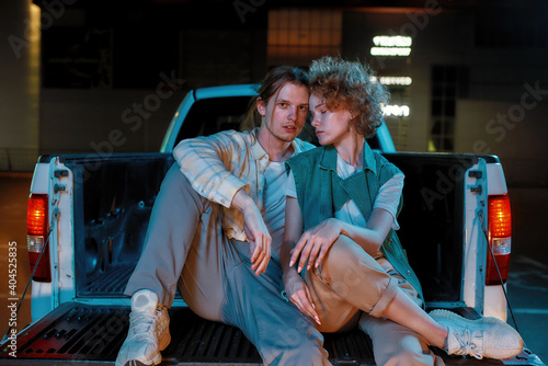 A group of two young good-looking white friends of different genders looking into a camera while sitting and posing in an opened car trunk outside on a parking site