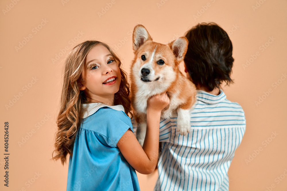 A boy stands with his back to the camera and a beautiful girl with curls in a dress and holds a cute funny dog on his shoulder isolated on a powdery peach background.Valentine's Day. A boy stands with his back to the camera and a beautiful girl with curls in a dress and holds a cute funny dog on his shoulder isolated on a powdery peach background.Valentine's Day.